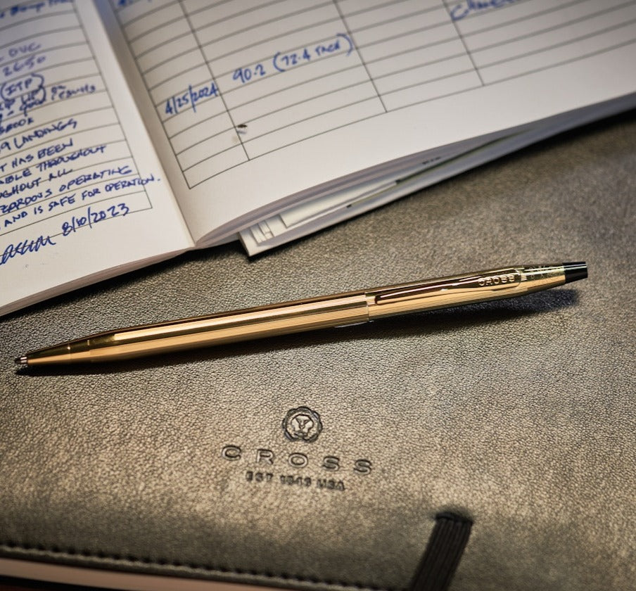 Gold pen on a leather surface with a notebook open in the background